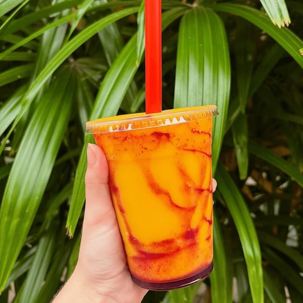 Hand holding a Big Island Bowls mango smoothie with red swirls and a red straw against green foliage.