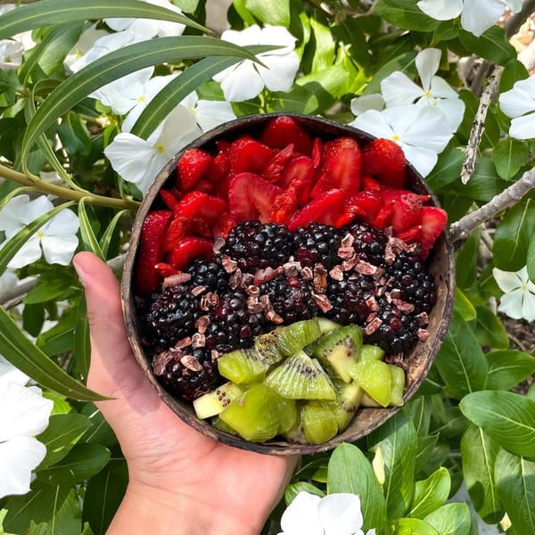 A hand holds a Big Island Bowls fruit bowl with strawberries, blackberries, and kiwi against white flowers.