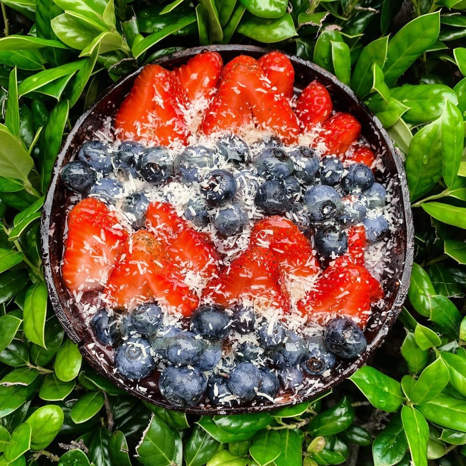 Big Island Bowls' acai bowl with strawberries, blueberries, and shredded coconut, surrounded by green leaves.