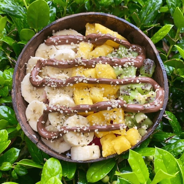 A Big Island Bowls acai bowl in a coconut shell, topped with banana, pineapple, kiwi, chocolate, and seeds.