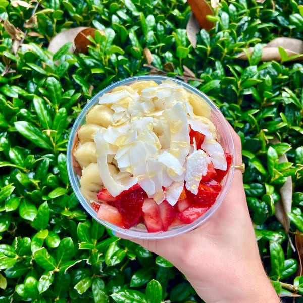 A hand holds a Big Island Bowls acai bowl topped with bananas, strawberries, and coconut flakes.