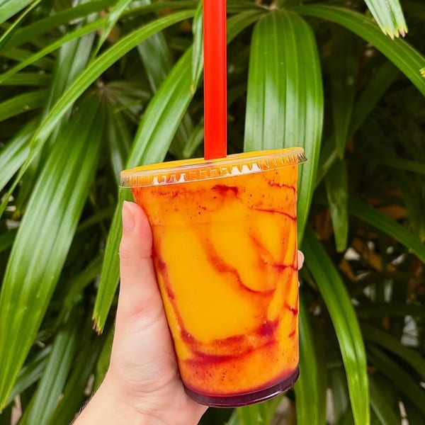 Hand holding a Big Island Bowls mango smoothie with red swirls and a red straw, against green foliage.