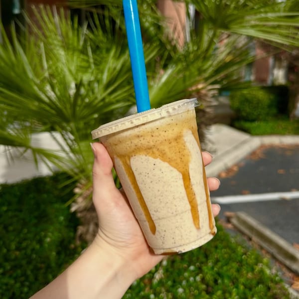 Hand holding a Big Island Bowls smoothie with caramel drizzle and a blue straw, outdoors.