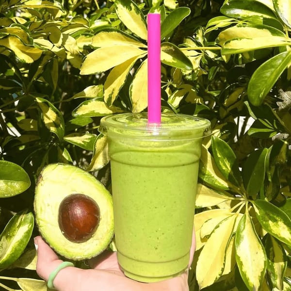 A hand holds a green smoothie with a pink straw and a halved avocado against lush foliage.